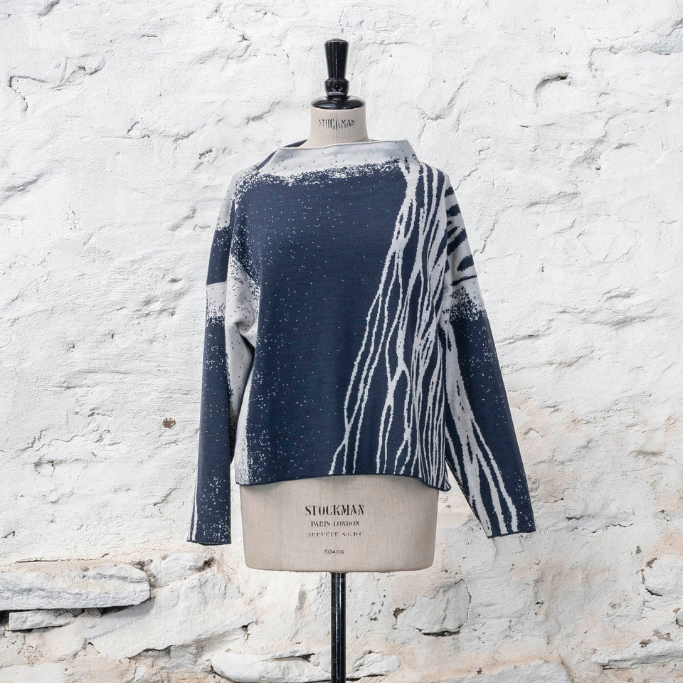 Contemporary jumper on a vintage mannequin against a rustic whitewashed wall, a finely knitted merino jumper in double faced jacquard. A linear design flows in waves down one side of the jumper - here on the front off-white against deep midnight blue (the jumper has been turned around). There are areas of white with speckle and blue areas with white speckle. The jumper is shown from the front and has a stand up neckline.