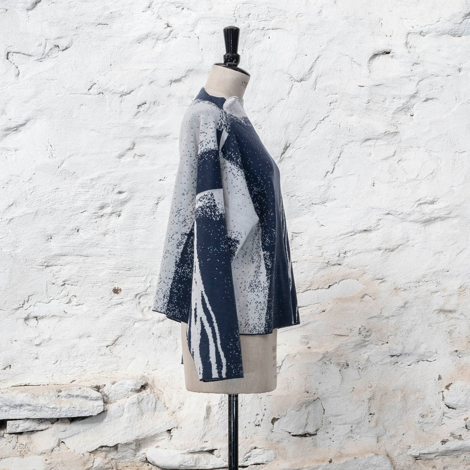 on a vintage mannequin against a rustic whitewashed wall, a finely knitted merino jumper in double faced jacquard. A linear design flows in waves down one side of the jumper - here on the front off-white against  deep midnight blue (the jumper has been turned around). There are areas of white with speckle and blue areas with white speckle.  The jumper  is shown from the right and has a stand up neckline.