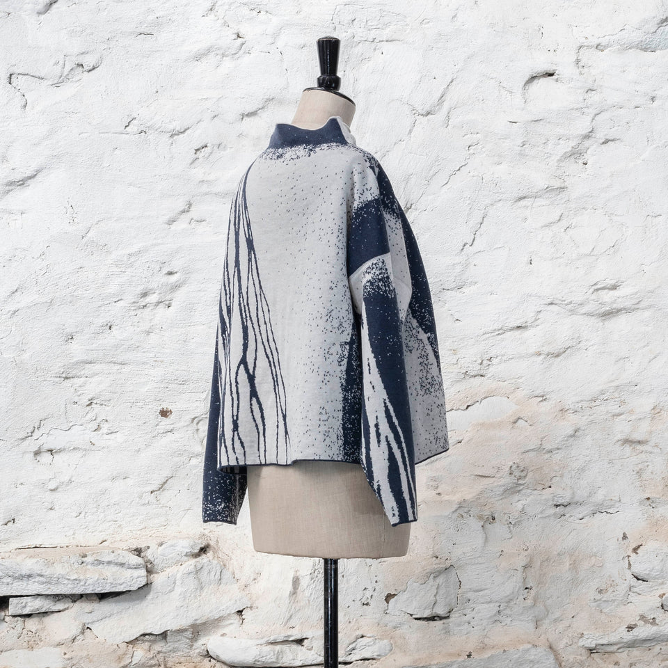 on a vintage mannequin against a rustic whitewashed wall, a finely knitted merino jumper in double faced jacquard. A linear design flows in waves down one side of the jumper - here on the back left shoulder in deep midnight blue against off white. There are areas of white with speckle and blue areas with white speckle. The jumper is shown from the back left at a three quarter angle and has a stand up neckline.
