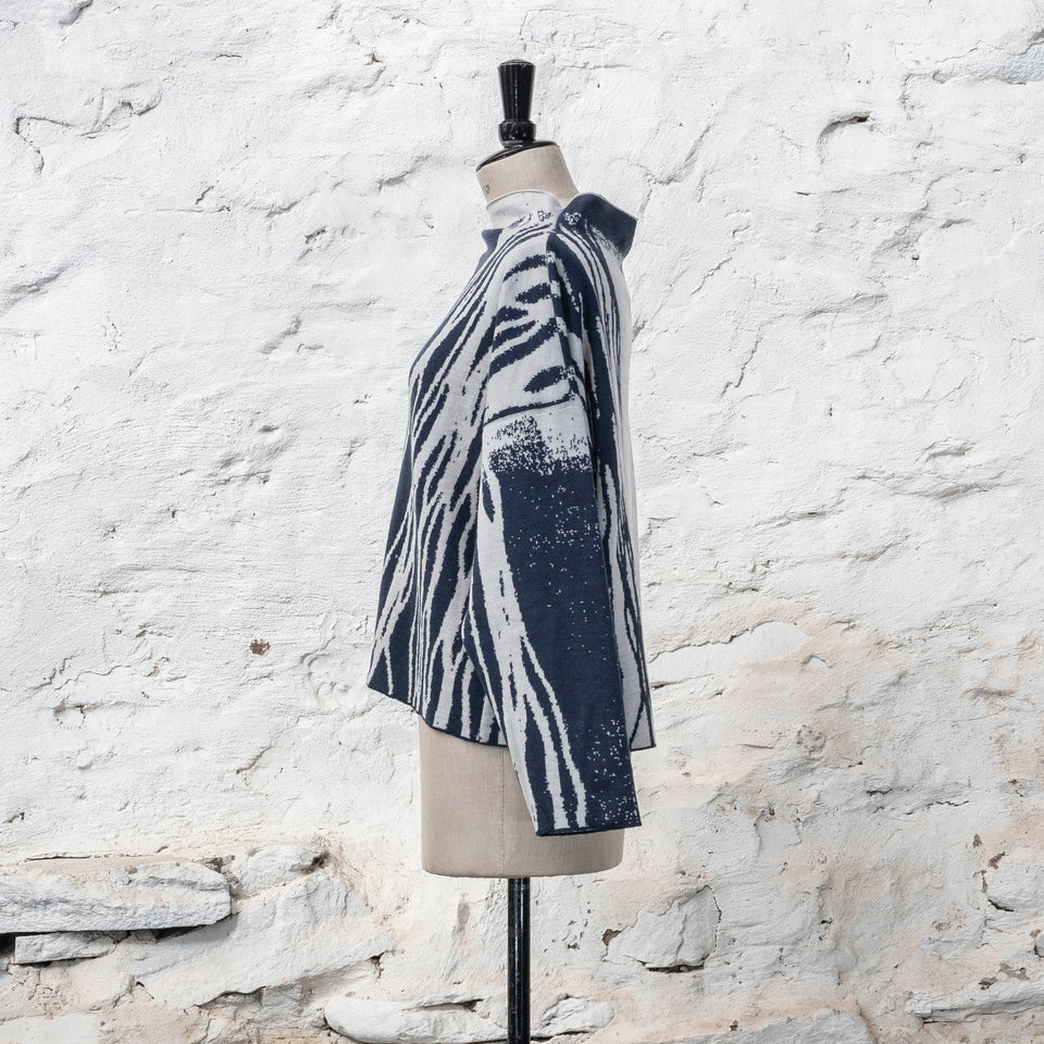 on a vintage mannequin against a rustic whitewashed wall, a finely knitted merino jumper in double faced jacquard. A linear design flows in waves down one side of the jumper - here on the back left shoulder in deep midnight blue against off white. There are areas of white with speckle and blue areas with white speckle. The jumper is shown from the left and has a stand up neckline