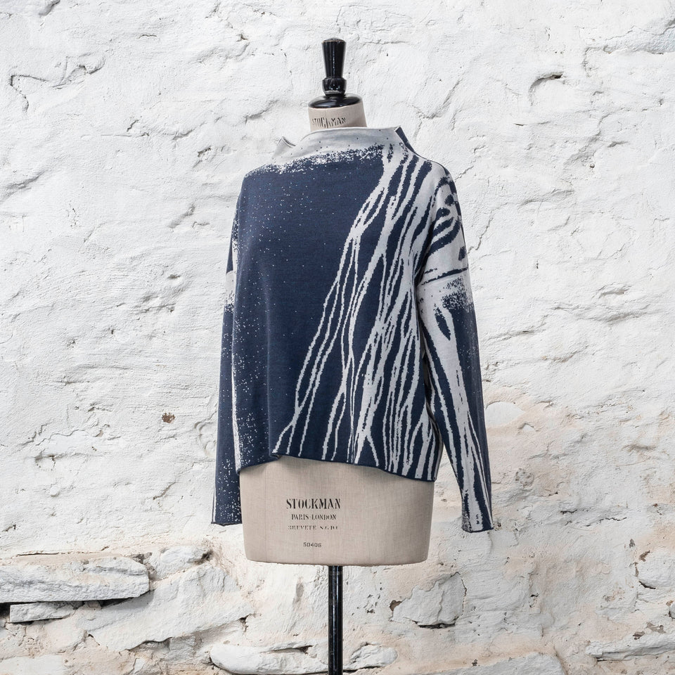 on a vintage mannequin against a rustic whitewashed wall, a finely knitted merino jumper in double faced jacquard. A linear design flows in waves down one side of the jumper - here on the front left shoulder in off white against deep midnight blue. There are areas of white with speckle and blue areas with white speckle. The jumper is shown from the front and has a stand up neckline