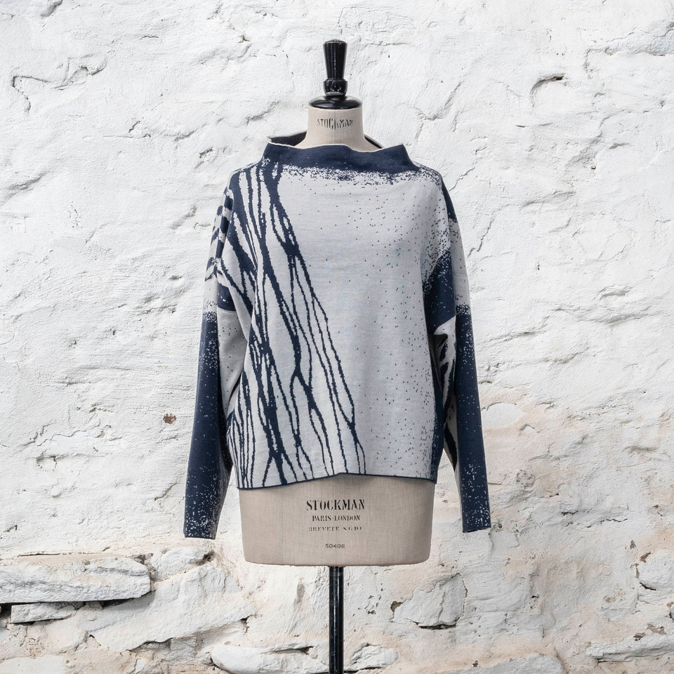 Graphic sweater on a stockman vintage mannequin. Shown with a rustic whitewashed wall as a background, a finely knitted merino jumper in double faced jacquard. A linear design flows in waves down one side of the jumper - here on the front in a deep midnight blue against off white. There are areas of white with speckle and blue areas with white speckle. The jumper is shown from the front and has a stand up neckline.