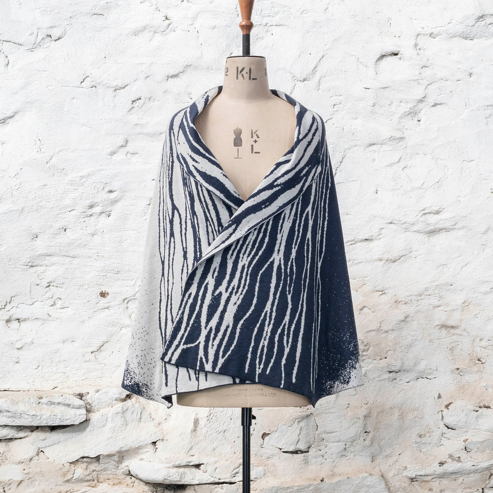 A midnight blue and off white knitted open cape in fine merino yarn. the pattern has fluid travelling likes either in blue on white or reversed out in white on blue. Shown from the front with linear panels just overlapping.