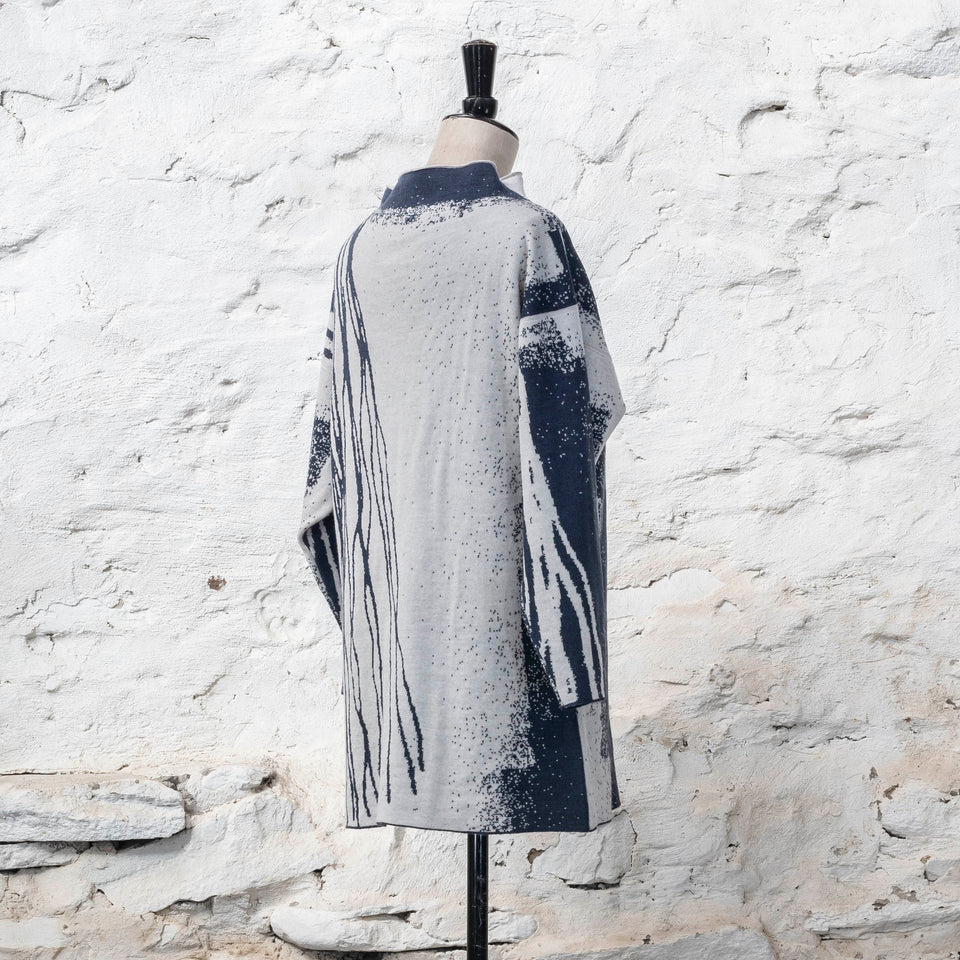 On a vintage mannequin, shown against a rustic whitewashed wall, a longline tunic jumper finely knitted in midnight blue and off white. Shown from the back where the pattern is predominantly white with blue lines travelling from the right shoulder down to the bottom of the garment. The tunic has a stand-up neck
