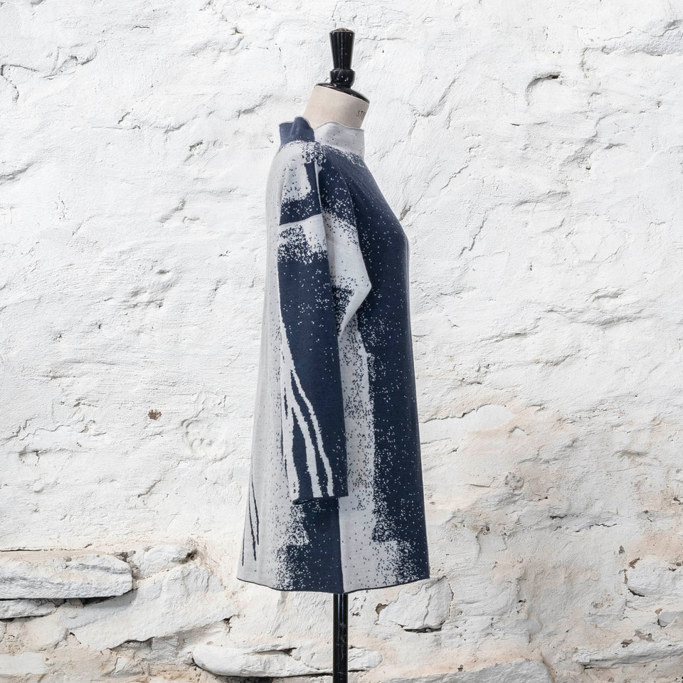 On a vintage mannequin, shown against a rustic whitewashed wall, a longline tunic jumper finely knitted in midnight blue and off white. Shown from the front where the pattern is predominantly white with blue lines travelling from the right shoulder down to the bottom of the garment. The tunic has a stand-up neck and is shown from the right side