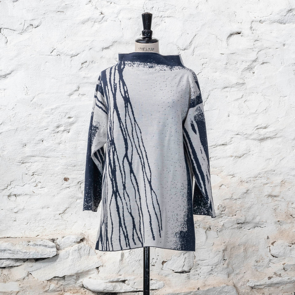 On a vintage mannequin, shown against a rustic whitewashed wall, a longline tunic jumper finely knitted in midnight blue and off white. Shown from the front where the pattern is predominantly white with blue lines travelling from the right shoulder down to the bottom of the garment. The tunic has a stand-up neck