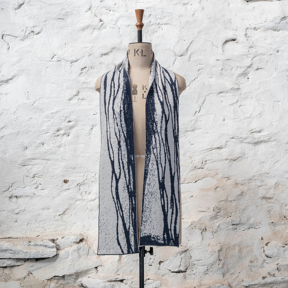 On a vintage mannequin shown against a rusting whitewashed wall, a midnight blue and off white finely knitted scarf in reverse jacquard. The pattern is linear wavs of line and speckle, moving along the scarf. Shown untied, open around the neck.