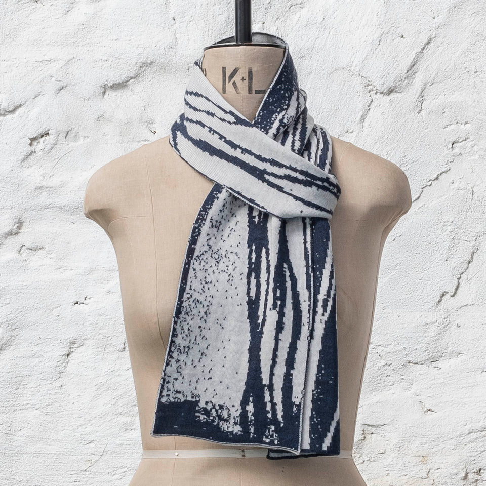 On a vintage mannequin shown against a rusting whitewashed wall, a midnight blue and off white finely knitted scarf in reverse jacquard. The pattern is linear wavs of line and speckle, moving along the scarf. Shown tied in a loop knot.