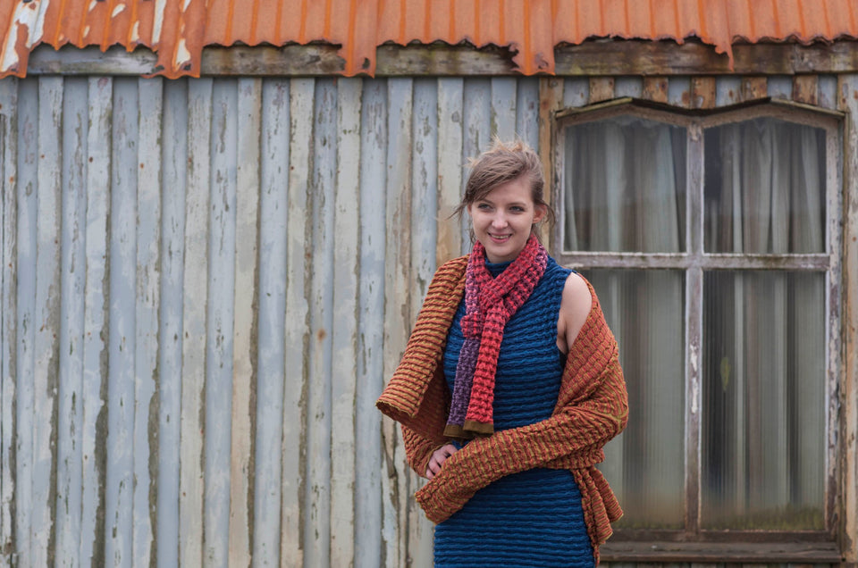 Model wears a rigg scarf, dress and jacket. The rigg textile is ridged and knitted in coloured stripes. Scarves have blocks of colour runing through the length