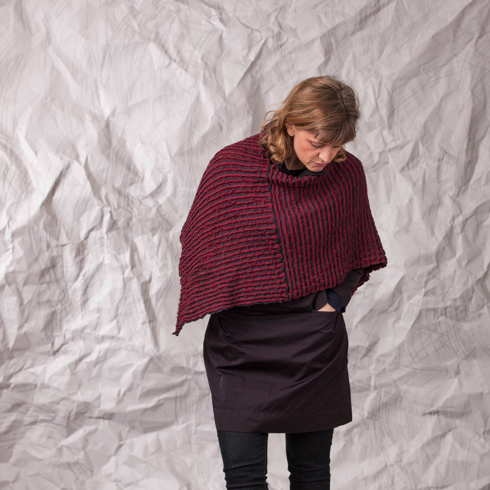 Model wears rigg cape. The textile is ridged and with stripes of colour knitted in. Shown here in a dark red and navy