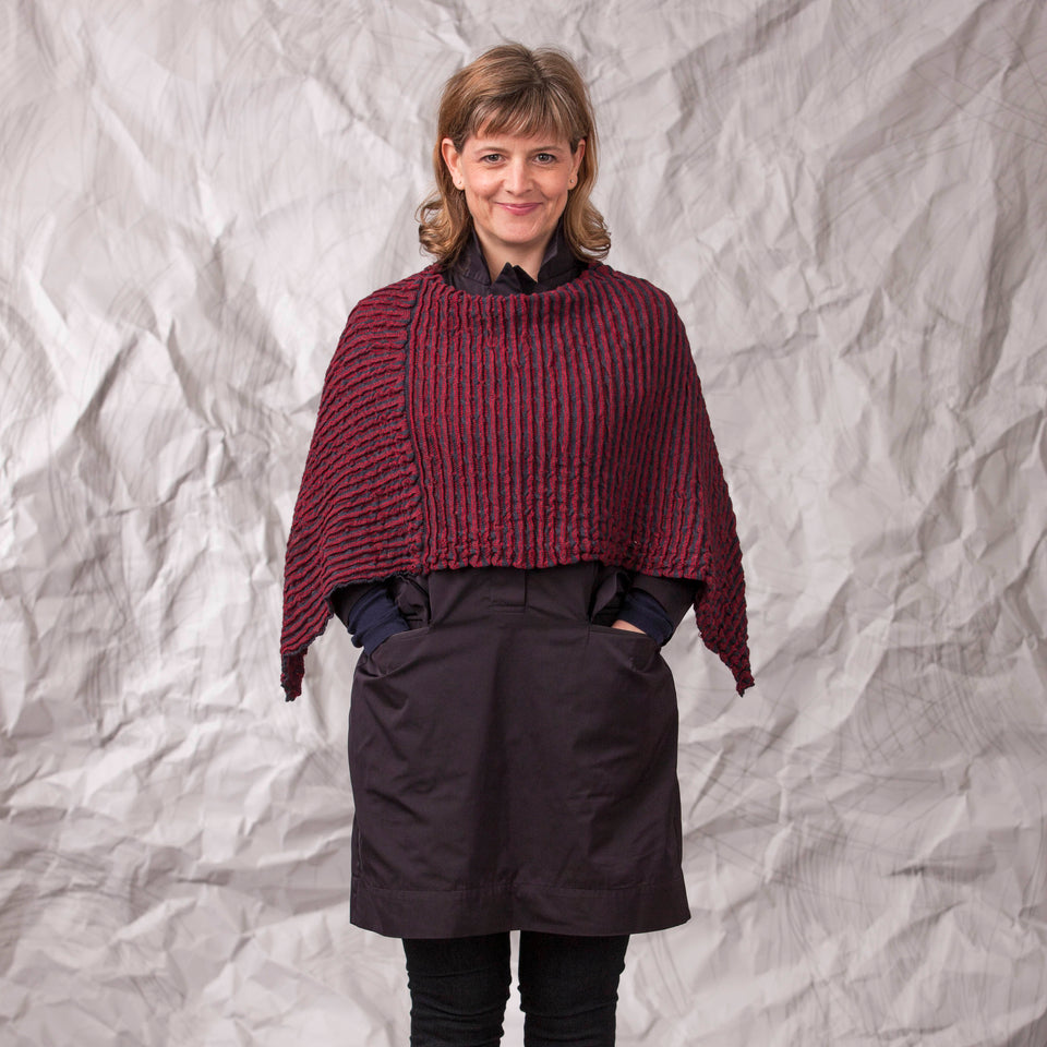 Model wears rigg cape. The textile is ridged and with stripes of colour knitted in. Shown here in a dark red and navy