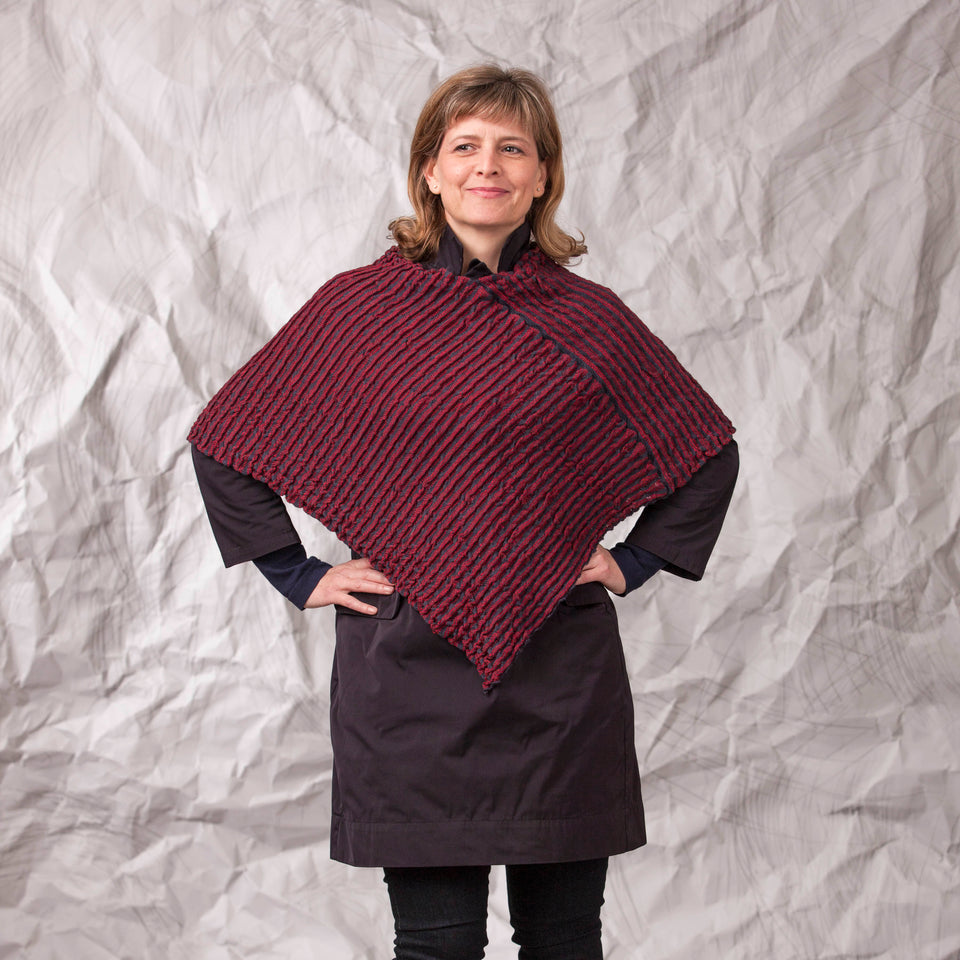 Model wears rigg cape. The textile is ridged and with stripes of colour knitted in. Shown here in a dark red and navy