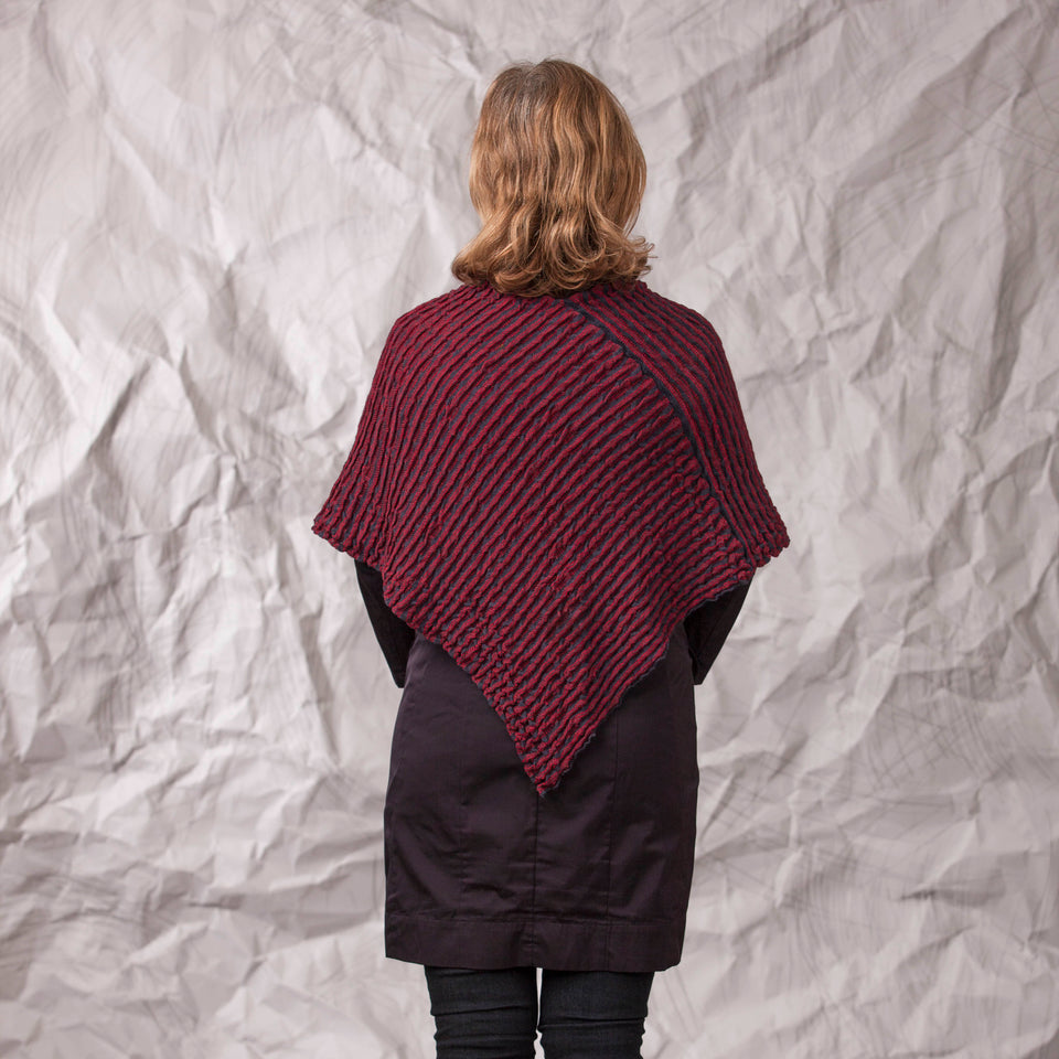 Model wears rigg cape/poncho. The textile is ridged and with stripes of colour knitted in. Shown here in a dark red and navy. Back view