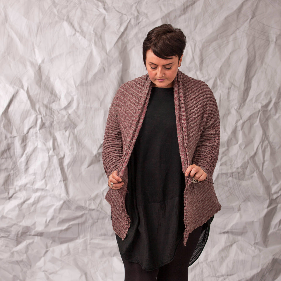 Model wears a longline knitted contemporary jacked in ridged fabric, in warm beige and cherry. Front view, holding the jacket slightly open.