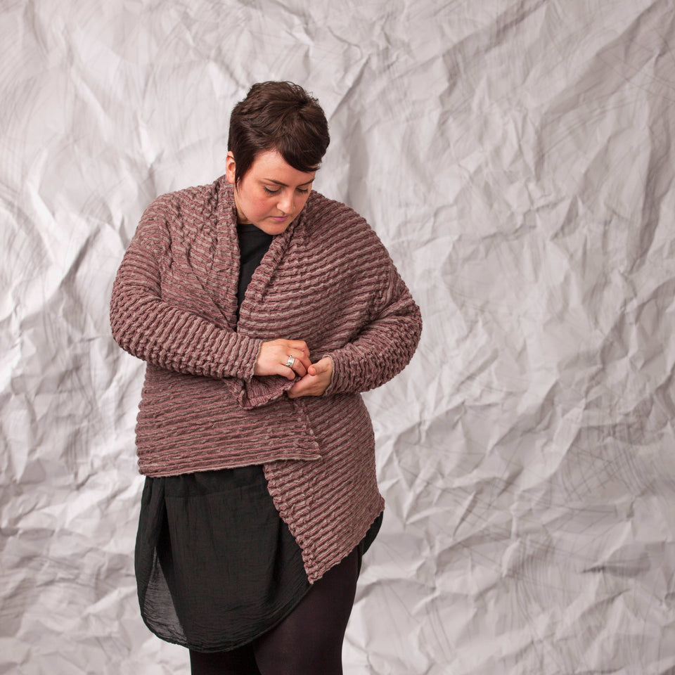 Model wears a longline knitted contemporary jacked in ridged fabric, in warm beige and cherry. Front view, with model pinning the jacket