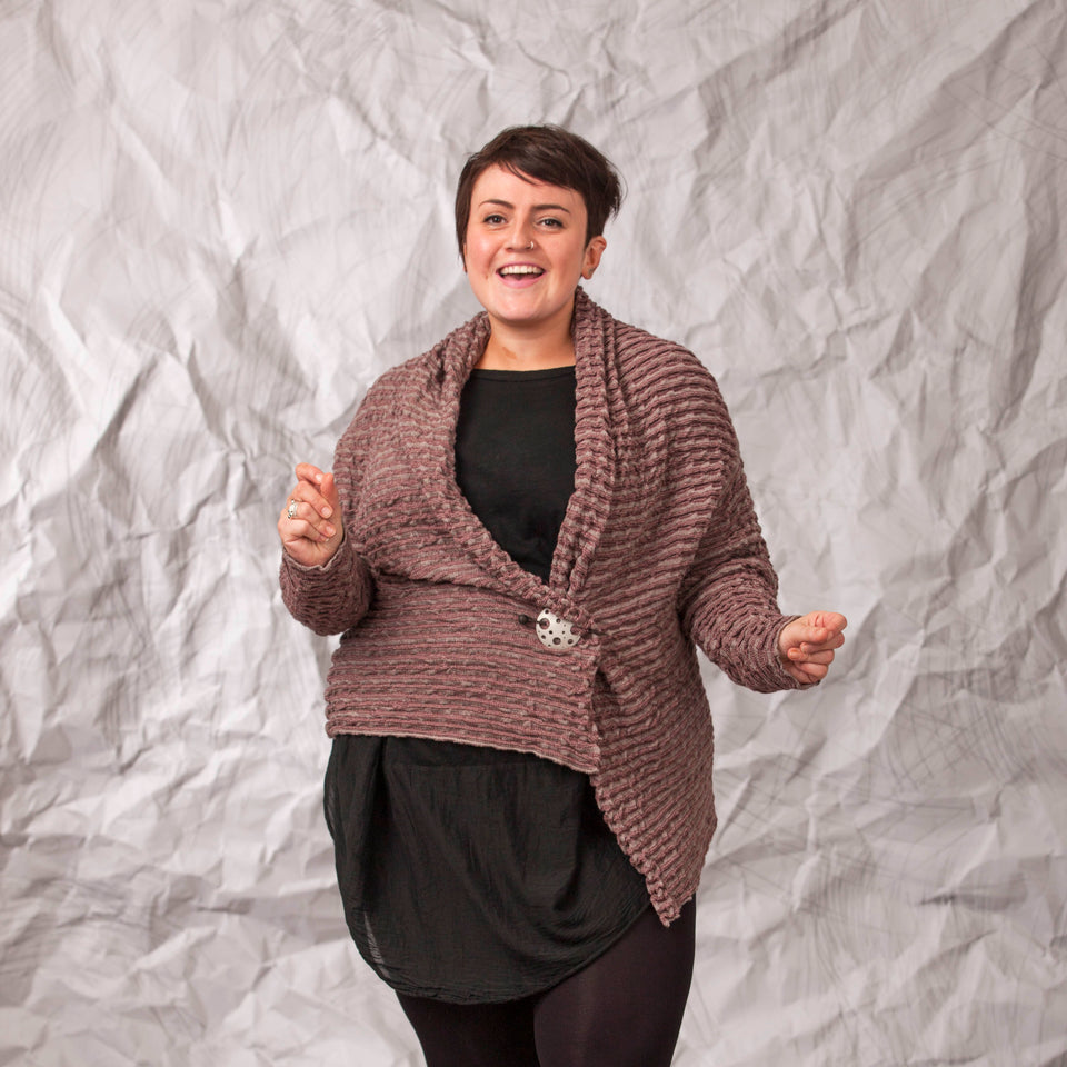 Model wears a longline knitted contemporary jacked in ridged fabric, in warm beige and cherry. Front view, with the jacket pinned shut