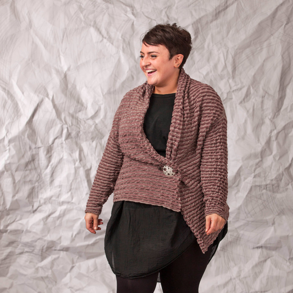 Model wears a longline knitted contemporary jacked in ridged fabric, in warm beige and cherry. Three-quarter view, with the jacket pinned shut