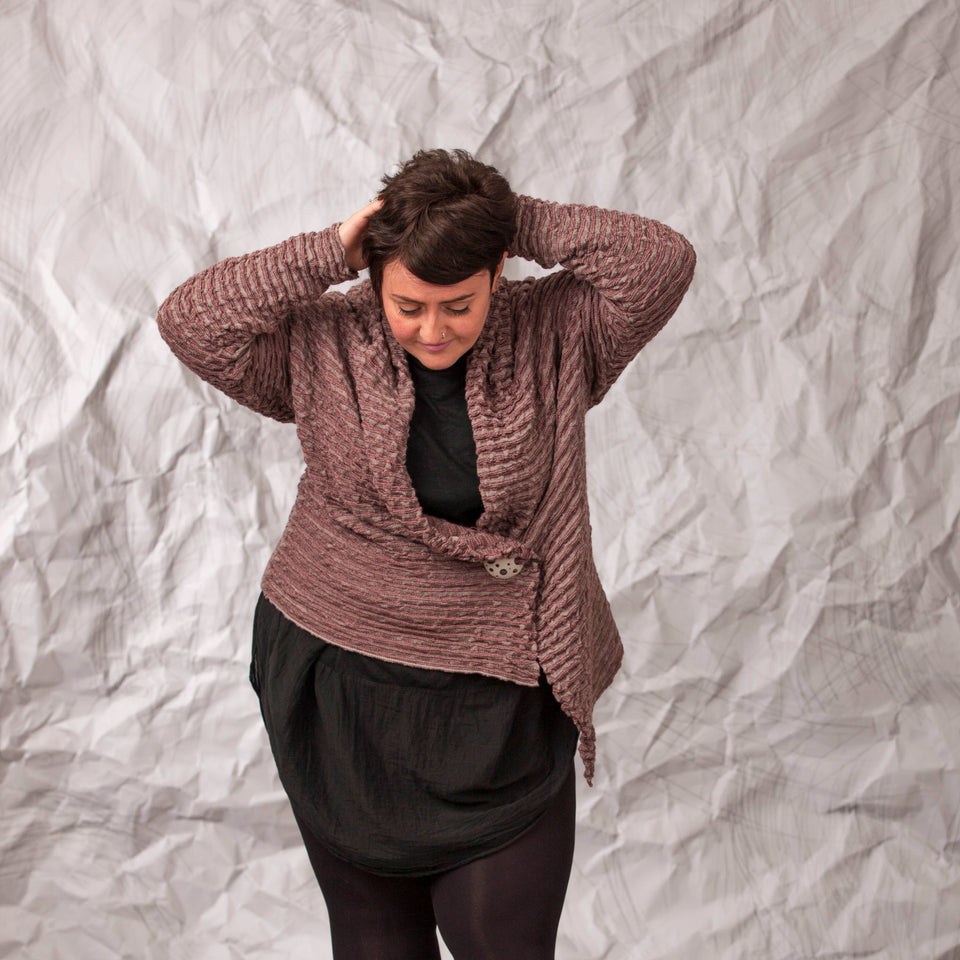 Model wears a longline knitted contemporary jacked in ridged fabric, in warm beige and cherry. Front view, with the jacket pinned shut and hands on head