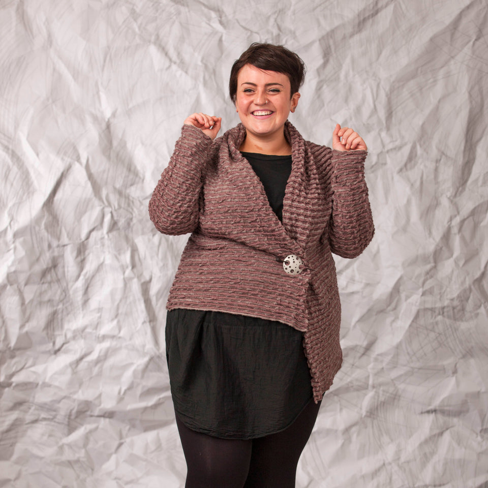 Model wears a longline knitted contemporary jacked in ridged fabric, in warm beige and cherry. Front view, with the jacket pinned shut