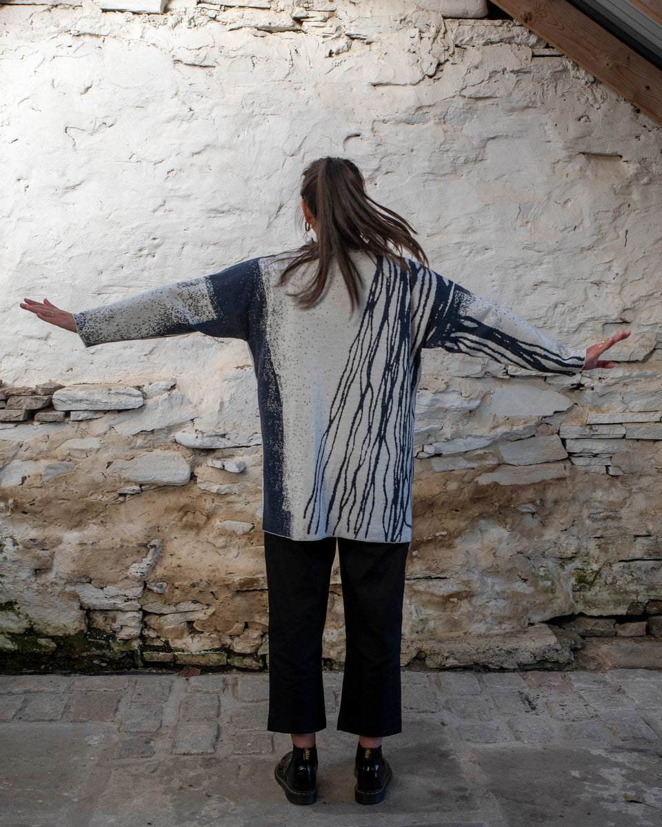 A woman with long dark hair and olive skin looks towards the camera. She is wearing cream metal hoop earrings and her he hair tied back in a ponytail, which swishes to one side. She is wearing a longline tunic jumper in navy and off white. The design is abstract and linera with large areas of speckled pattern. She is also wearing black trousers and is standing in a stone, whitewashed shed in hoswick Shetland.