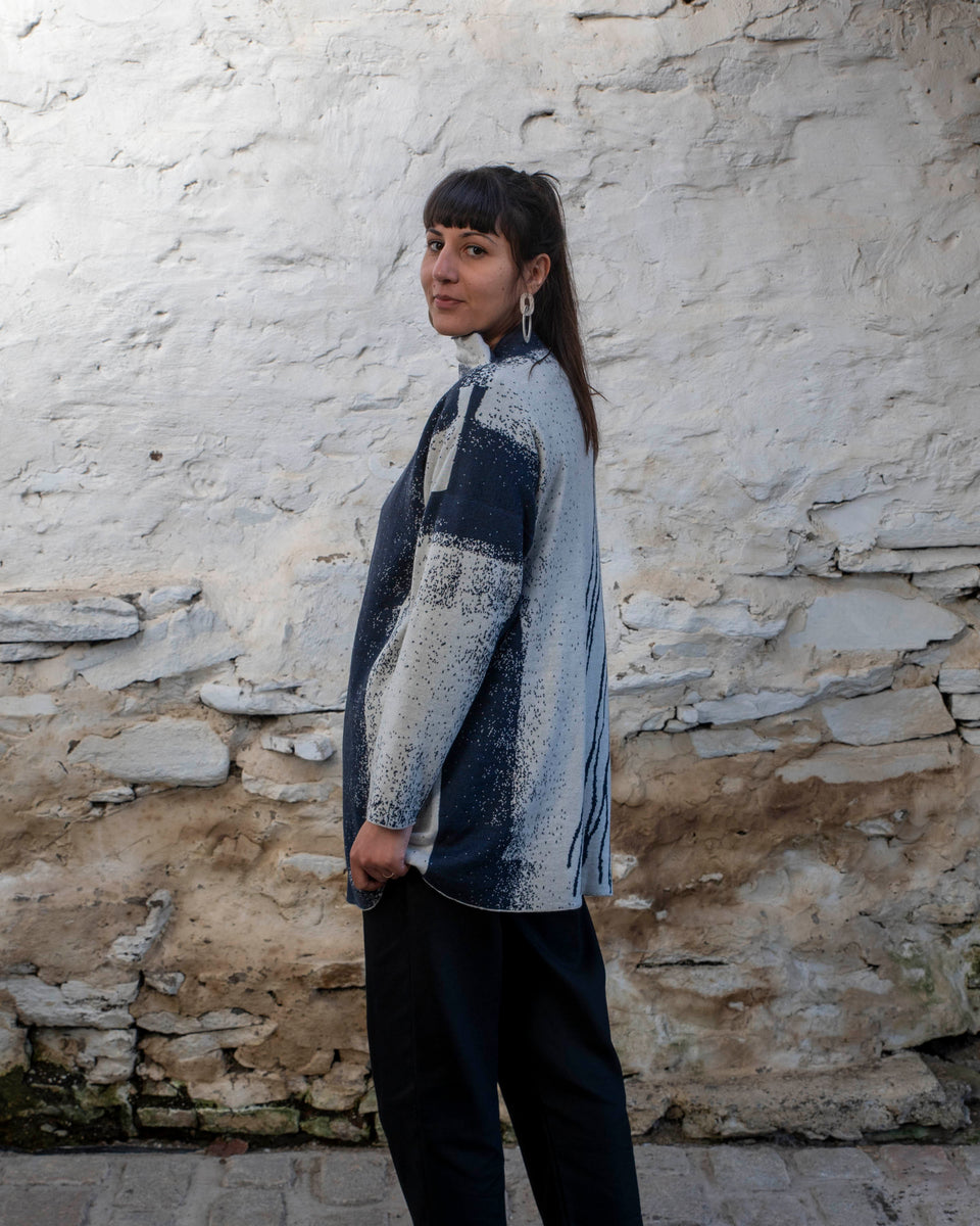 A woman with long dark hair and olive skin looks towards the camera. She is wearing cream metal hoop earrings and her he hair tied back in a ponytail, which swishes to one side. She is wearing a longline tunic jumper in navy and off white. The design is abstract and linera with large areas of speckled pattern. She is also wearing black trousers and is standing in a stone, whitewashed shed in hoswick Shetland.