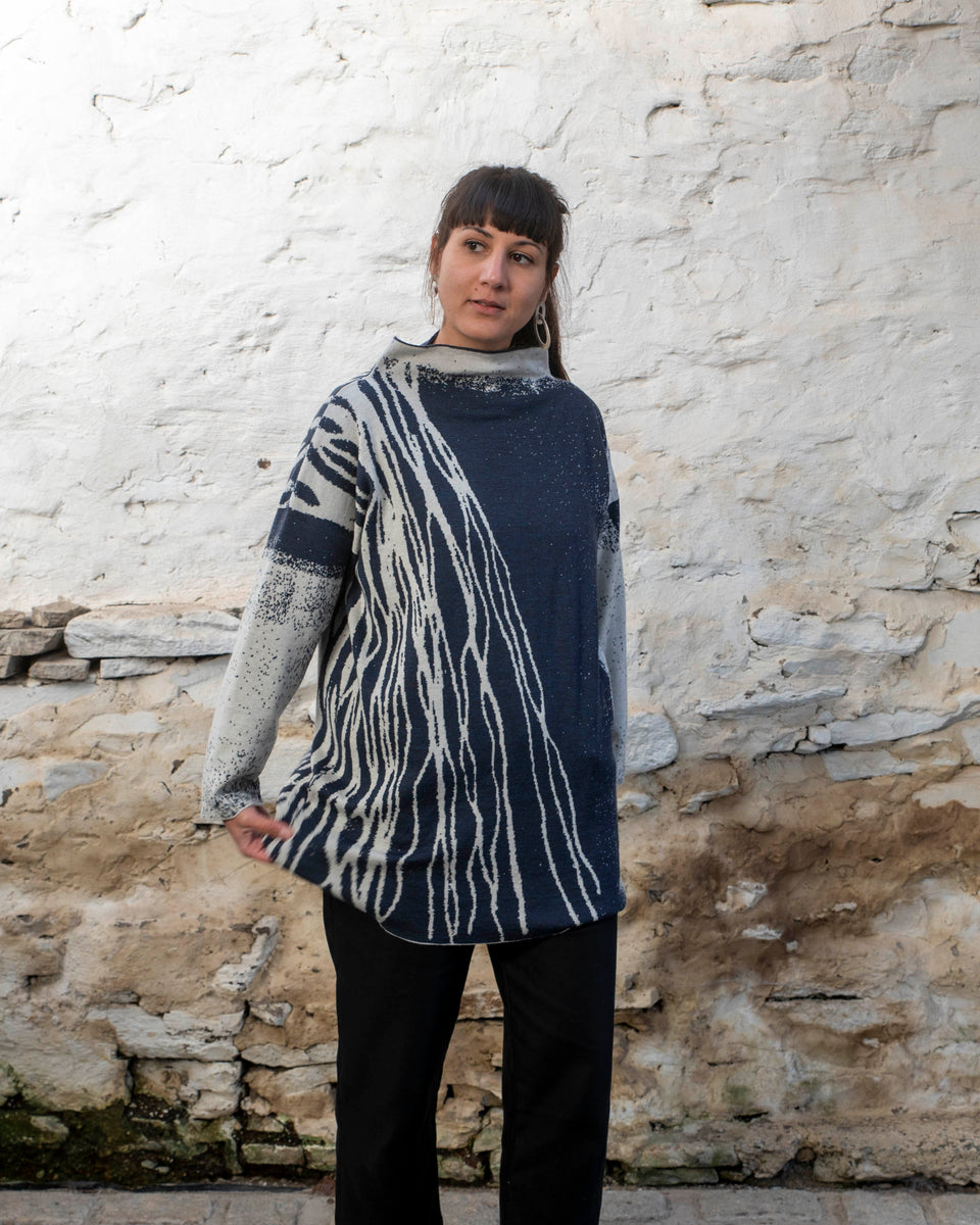 A woman with long dark hair and olive skin looks towards the camera. She is wearing cream metal hoop earrings and her he hair tied back in a ponytail, which swishes to one side. She is wearing a longline tunic jumper in navy and off white. The design is abstract and linera with large areas of speckled pattern. She is also wearing black trousers and is standing in a stone, whitewashed shed in hoswick Shetland.