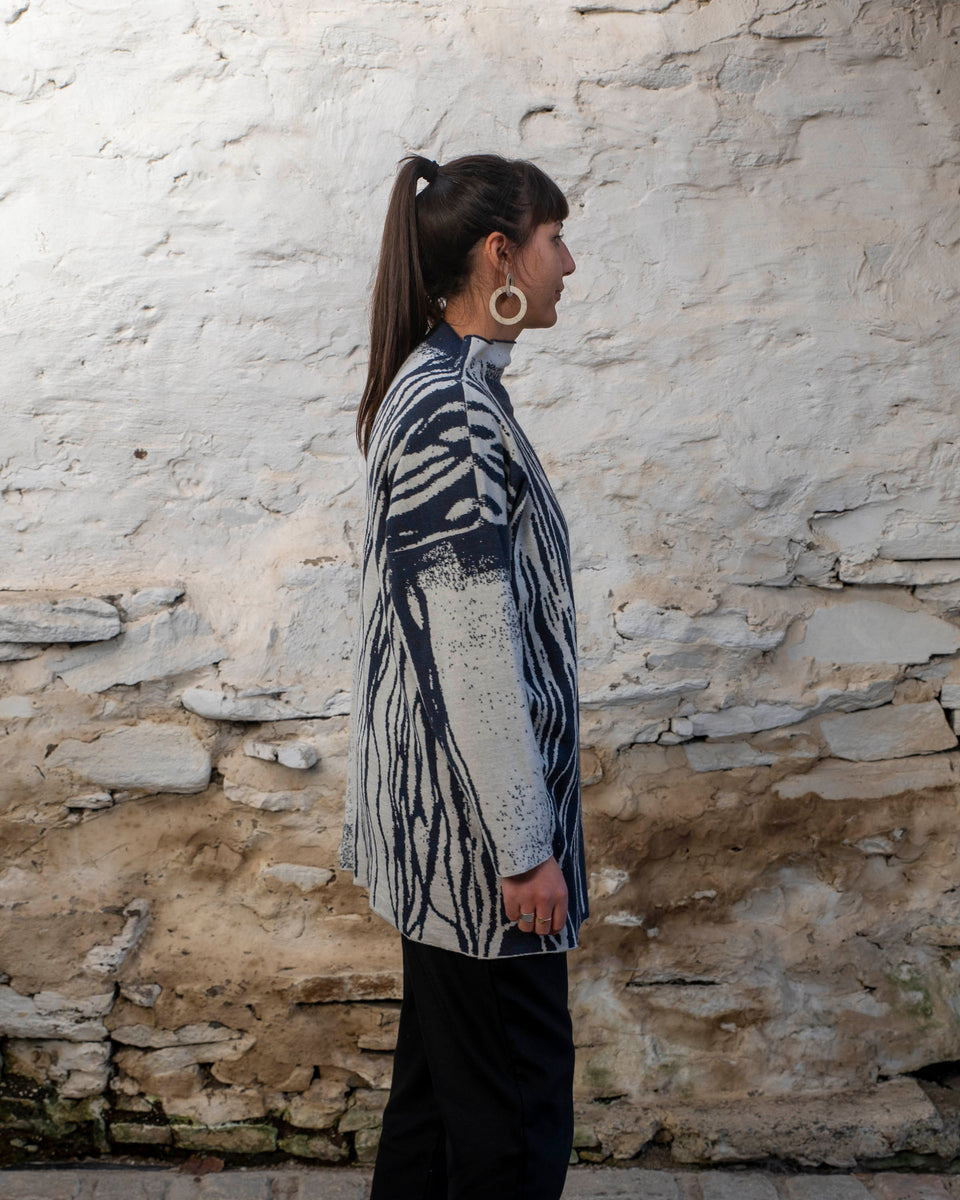 A woman with long dark hair and olive skin looks towards the camera. She is wearing cream metal hoop earrings and her he hair tied back in a ponytail, which swishes to one side. She is wearing a longline tunic jumper in navy and off white. The design is abstract and linera with large areas of speckled pattern. She is also wearing black trousers and is standing in a stone, whitewashed shed in hoswick Shetland.