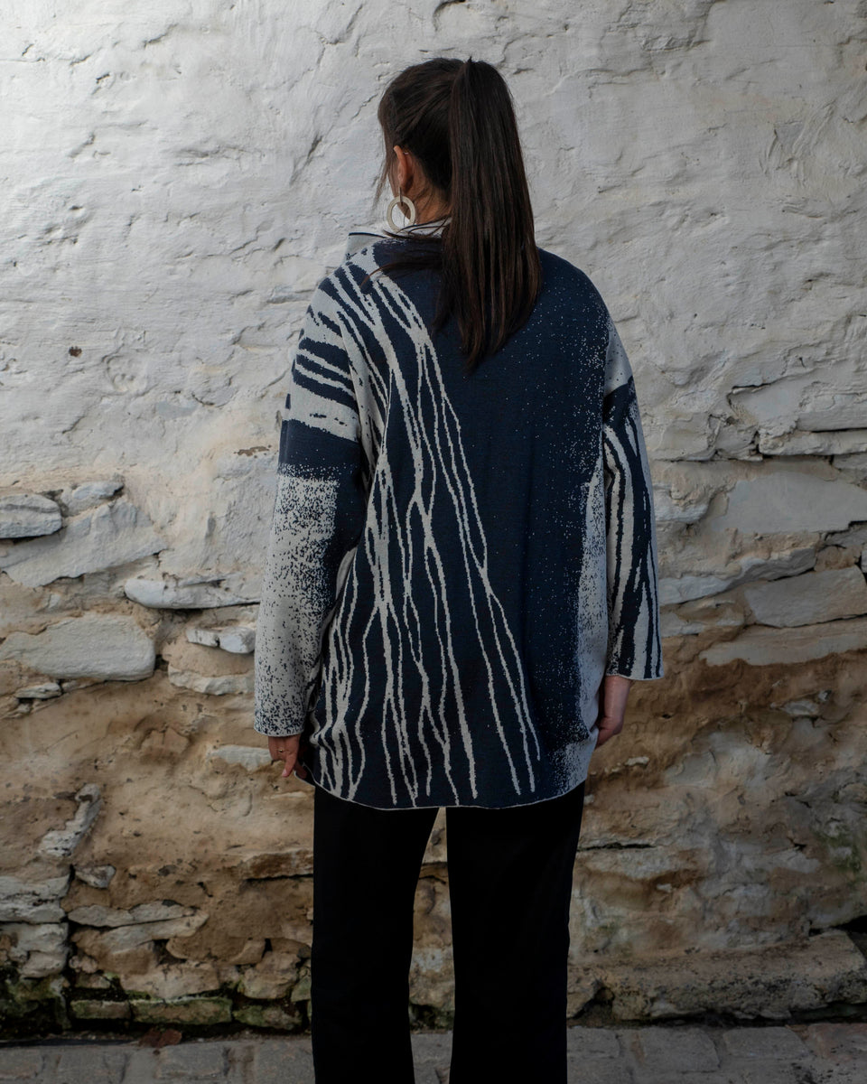 A woman with long dark hair and olive skin looks towards the camera. She is wearing cream metal hoop earrings and her he hair tied back in a ponytail, which swishes to one side. She is wearing a longline tunic jumper in navy and off white. The design is abstract and linera with large areas of speckled pattern. She is also wearing black trousers and is standing in a stone, whitewashed shed in hoswick Shetland.