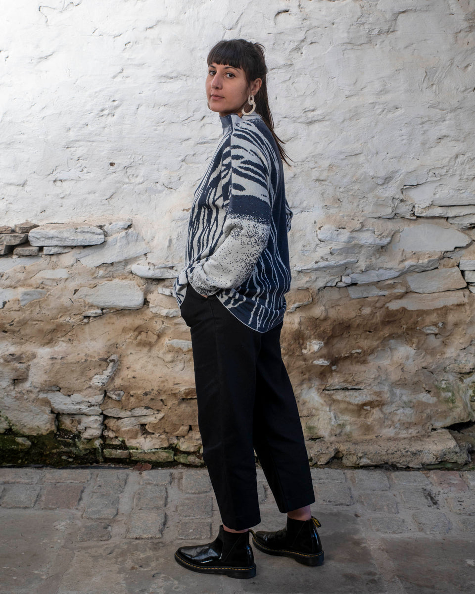 A woman with long dark hair and olive skin looks towards the camera. She is wearing cream metal hoop earrings and her he hair tied back in a ponytail, which swishes to one side. She is wearing a longline tunic jumper in navy and off white. The design is abstract and linera with large areas of speckled pattern. She is also wearing black trousers and is standing in a stone, whitewashed shed in hoswick Shetland.