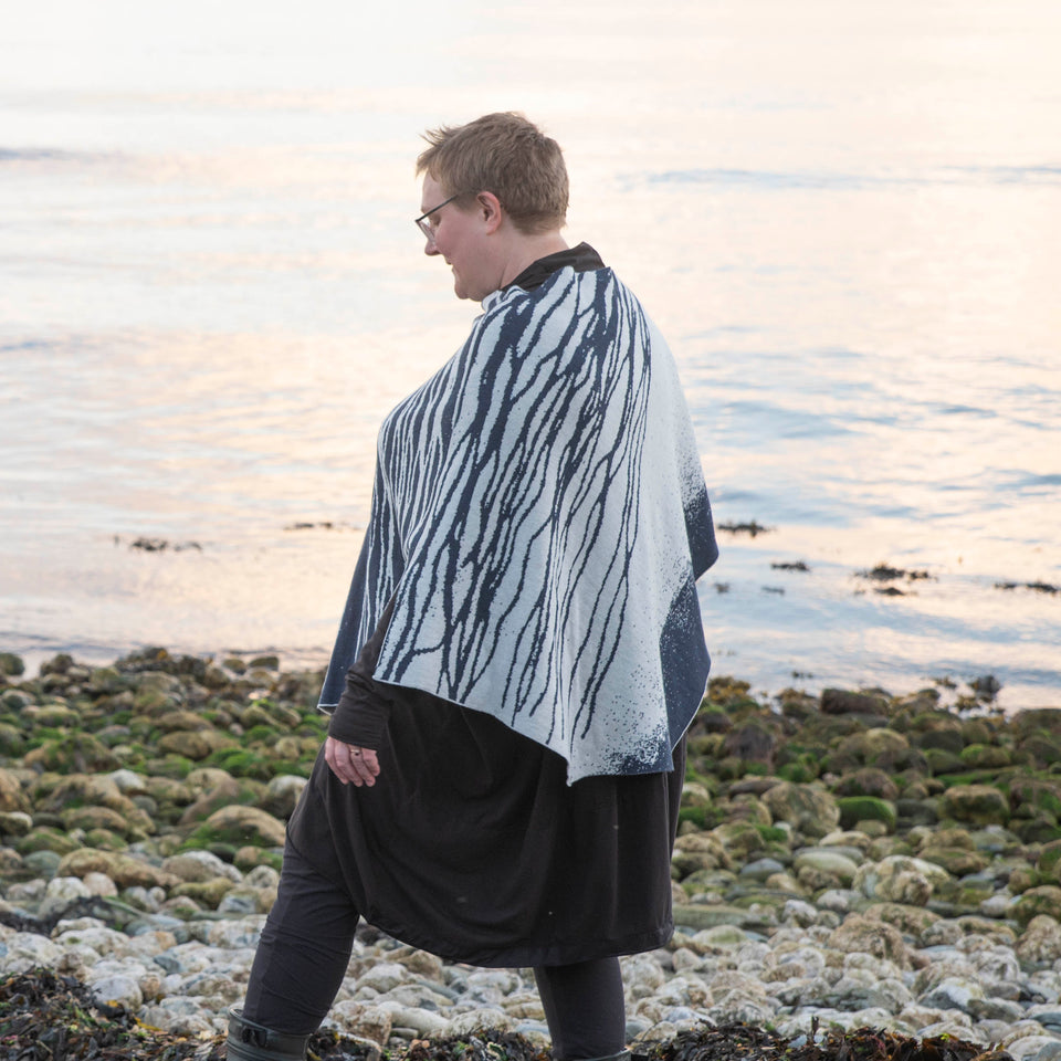 A white woman with short fair hair stands on a pebbly beach in Hoswick Shetland. She is wearing purplish clear spectacles, an off black rundholz tunic and leggings. Over this she wears a finely knitted cape by Nielanell. The design is abstract with flowing lines reminiscent of water rivulets on sand and is knitted in inky blue and off white reverse jacquard.