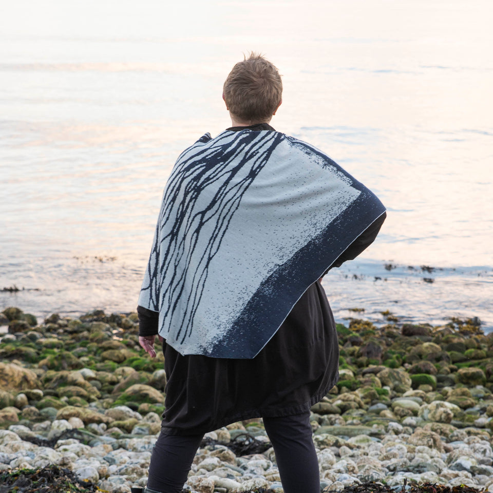 A white woman with short fair hair stands on a pebbly beach in Hoswick Shetland. She is wearing purplish clear spectacles, an off black rundholz tunic and leggings. Over this she wears a finely knitted cape by Nielanell. The design is abstract with flowing lines reminiscent of water rivulets on sand and is knitted in inky blue and off white reverse jacquard.