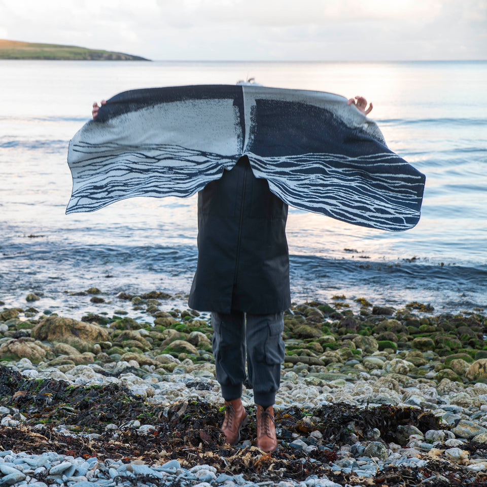 A white woman stands on the pebbly shore at Hoswick, Shetland. She is wearing a black 3/4 lenght coat, grey trousers with pockets on the sides and brown lace-up boots. She holds up a contemporary Shetland shawl which is knitted in midnight blue and off white with an abstract flowing lines pattern ressembling water rivulets on sand. Her head and shoulders are obscured by the shawl.
