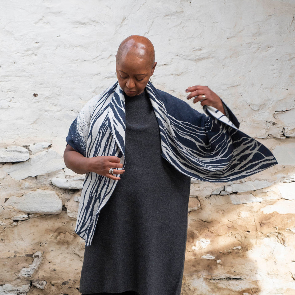 A black woman with a shaved head stands in a rustic, whitewashed stone shed. She is wearing a short sleeved, charcoal grey knitted dress. Over this she wears an inky blue and off-white wrap. The pattern is liner and abstract with flowing lines running downwards from her neck. This is the Shoormal design by Nielanell, which has echoes of the sand at the water's edge. She moves the wrap, throwing the left side out to reveal more pattern.
