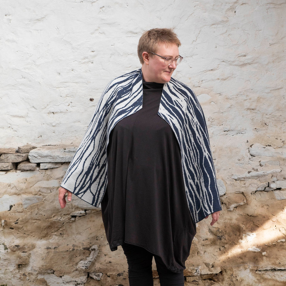 A white woman with short, fair hair stands front on to the camera with her head turned to her left, in a rustic, whitewashed Shetland stone shed. She is wearing spectacles with thin metal legs. Over an off-black tunic she wears a contemporary, finely knitted shawl. A pattern of flowing lines travel down her shoulders and arms arms. With her arms low at her sides the shawl looks somewhat like a cape.