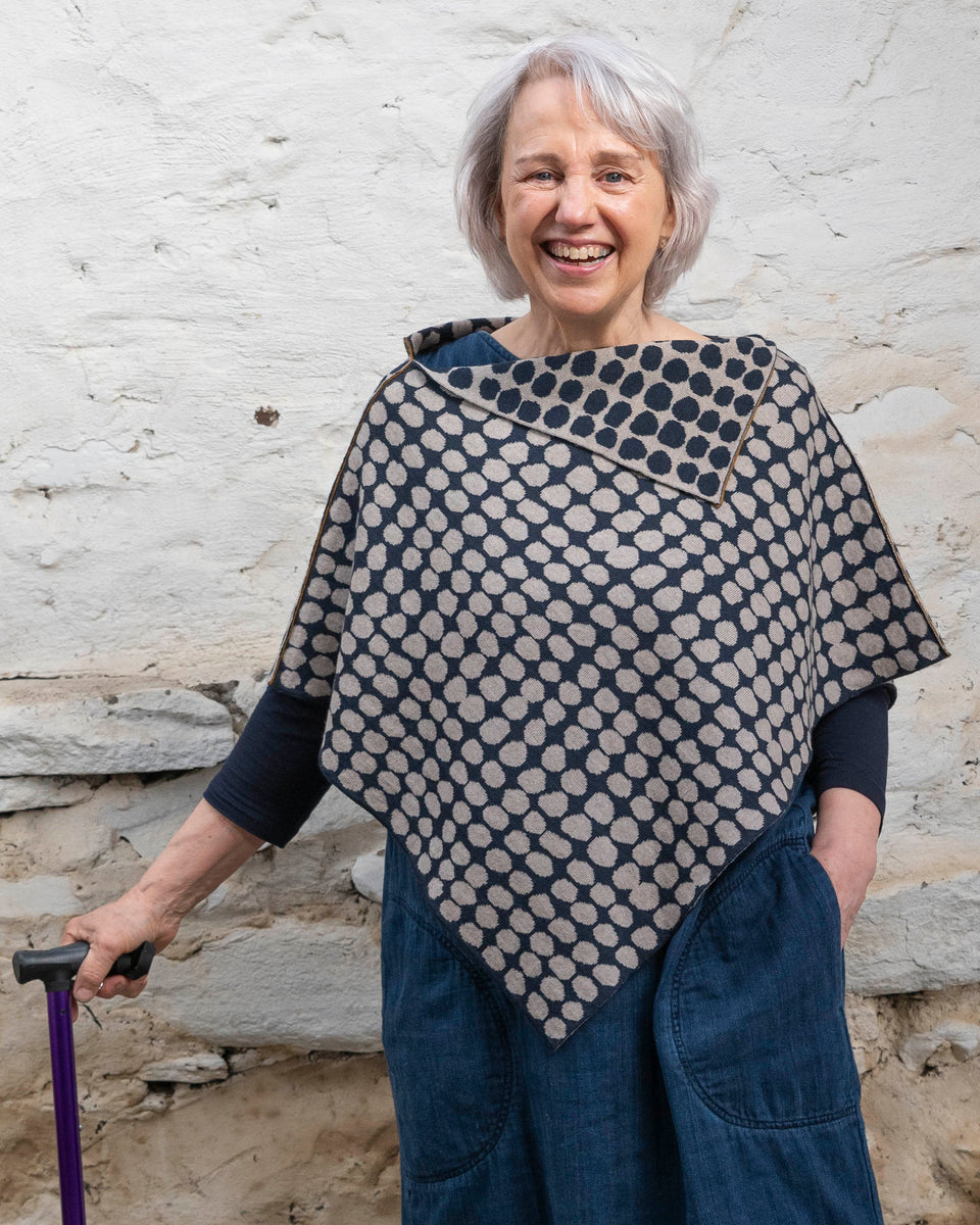 A white woman with jaw length grey hair stands in a rustic, whitewashed stone building. She has a purple walking stick in her right hand. She wears a finely knitted cape in navy and fawn in an irregular dot pattern, over a denim dress. 