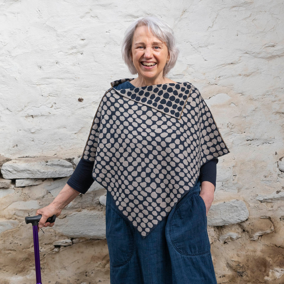 A white woman with jaw length grey hair stands in a rustic, whitewashed stone building. She has a purple walking stick in her right hand. She wears a finely knitted cape in navy and fawn in an irregular dot pattern, over a denim dress.