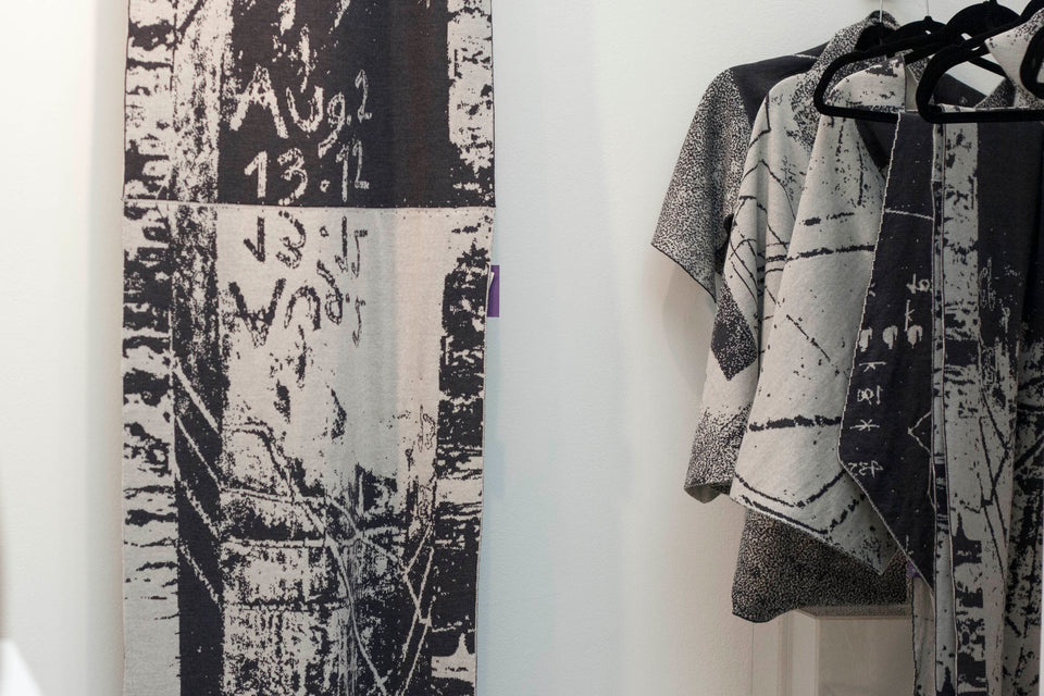 Knitted Byre wrap/shawl hanging alongside other pieces from the Byre collection. Abstract, graphic design in an asymmetric shape. Shown in Charcoal and stone white