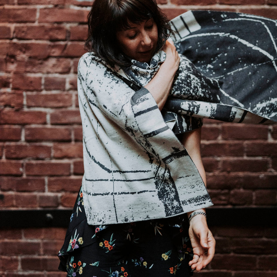 Model wears knitted Byre wrap/shawl. Abstract, graphic design in an asymmetric shape. Shown in Charcoal and stone white