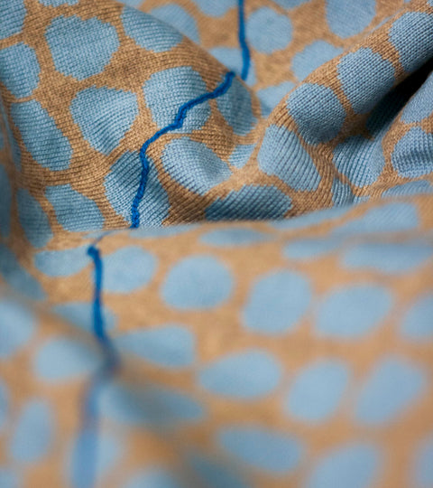 Detail of a jumper from the Nielanell Ebb-stanes collection, a collection of contemporary knitwear inspired by borders. A knitted blue line runs across a background of sky blue dots on ochre yellow. Knitted in extra fine merino