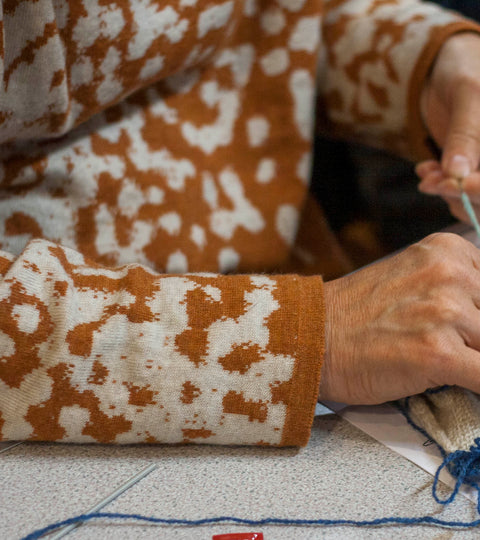 At Shetland Wool Week in a knitting class in Hoswick, a participant works with a swatch of knitting. She is wearing a Nielanell jumper in a mottled pattern of burnt orange and off white.