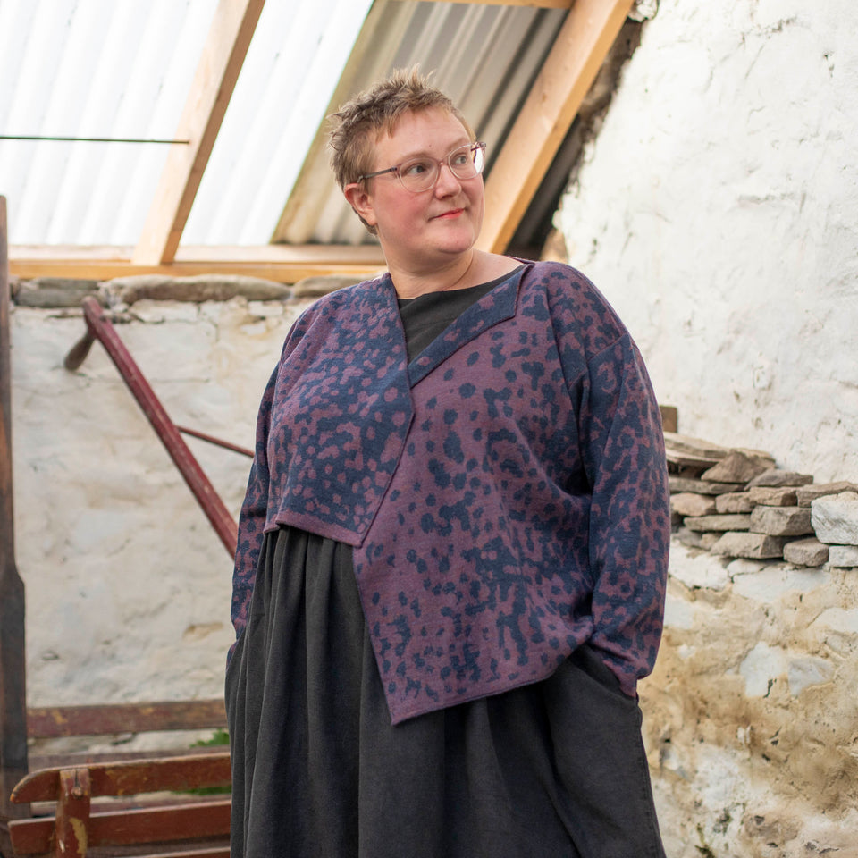 A white woman with short hair and clear framed spectacles wears a piece of contemporary Scottish knitwear - a knitted cardigan in animal print with blue and a soft purple over a black linen dress.