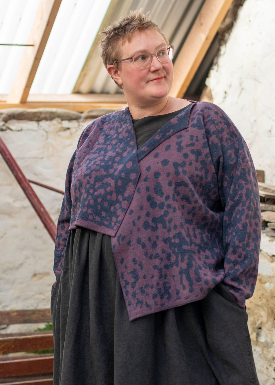 A white woman with short hair and clear framed spectacles wears a piece of contemporary Scottish knitwear - a knitted cardigan in animal print with blue and a soft purple over a black linen dress.