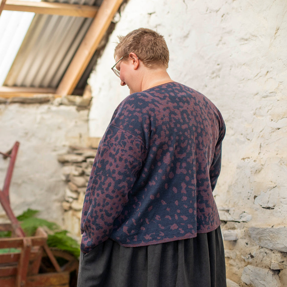 A white woman with short hair and clear framed spectacles wears a piece of contemporary Scottish knitwear - a knitted cardigan in animal print with blue and a soft purple over a black linen dress.