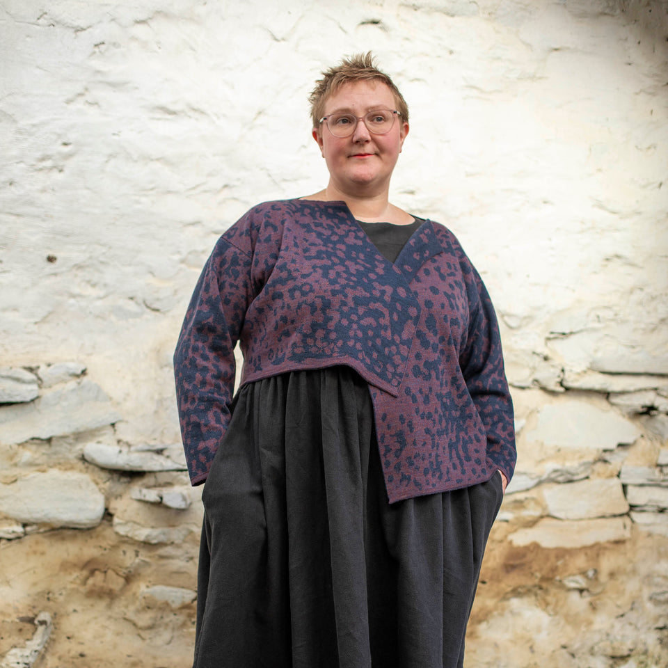 A white woman with short hair and clear framed spectacles wears a piece of contemporary Scottish knitwear - a knitted cardigan in animal print with blue and a soft purple over a black linen dress.