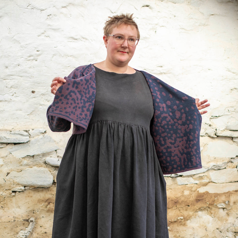 A white woman with short hair and clear framed spectacles wears a piece of contemporary Scottish knitwear - a knitted cardigan in animal print with blue and a soft purple over a black linen dress.