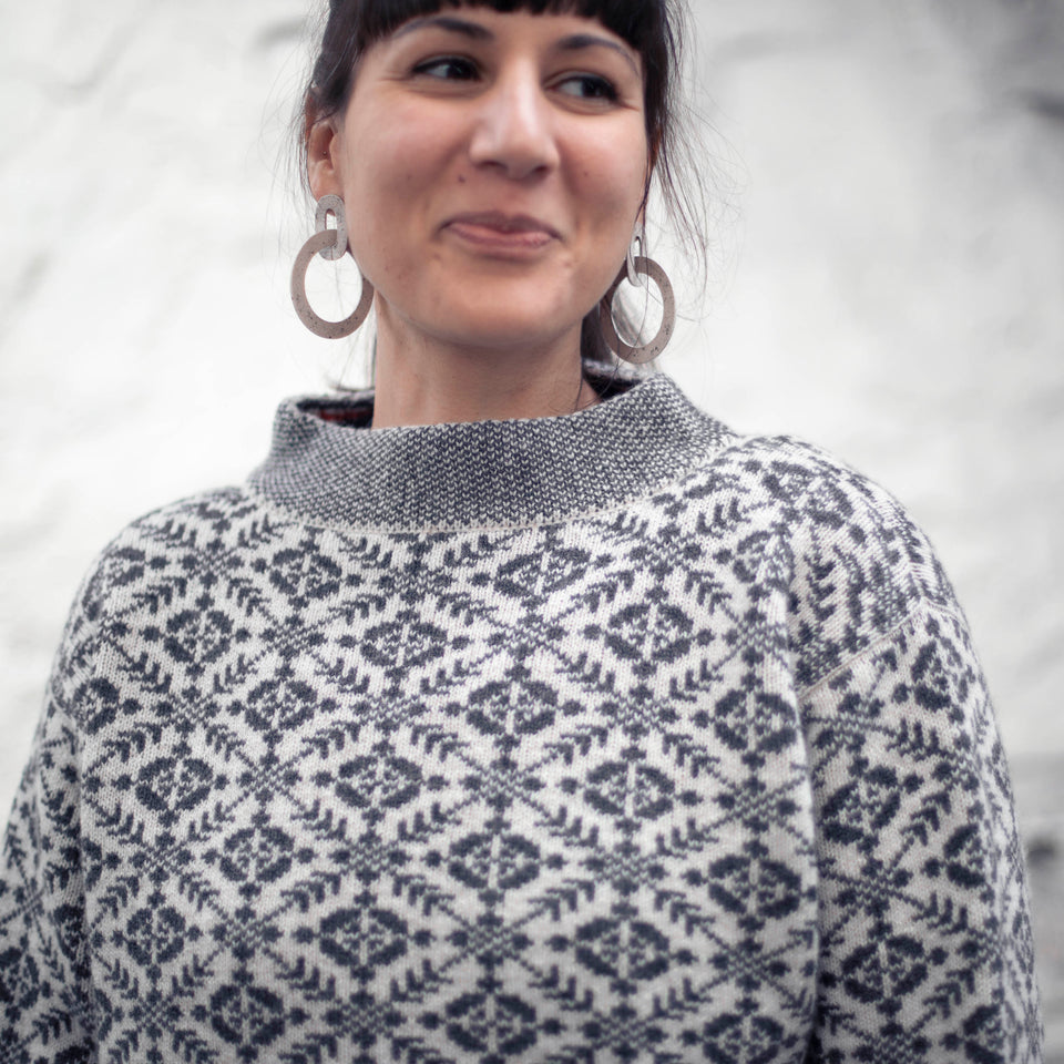 A woman with dark hair with a fringe and tied back into a pony tail wears a charcoal and white fair isle nielanell smookie jumper. It has an all over pattern and a stand up neck in bidseye stitch. She is smiling and looking away from the camera, to the side. A rustic, whitewashed stone wall is out of focus behind.