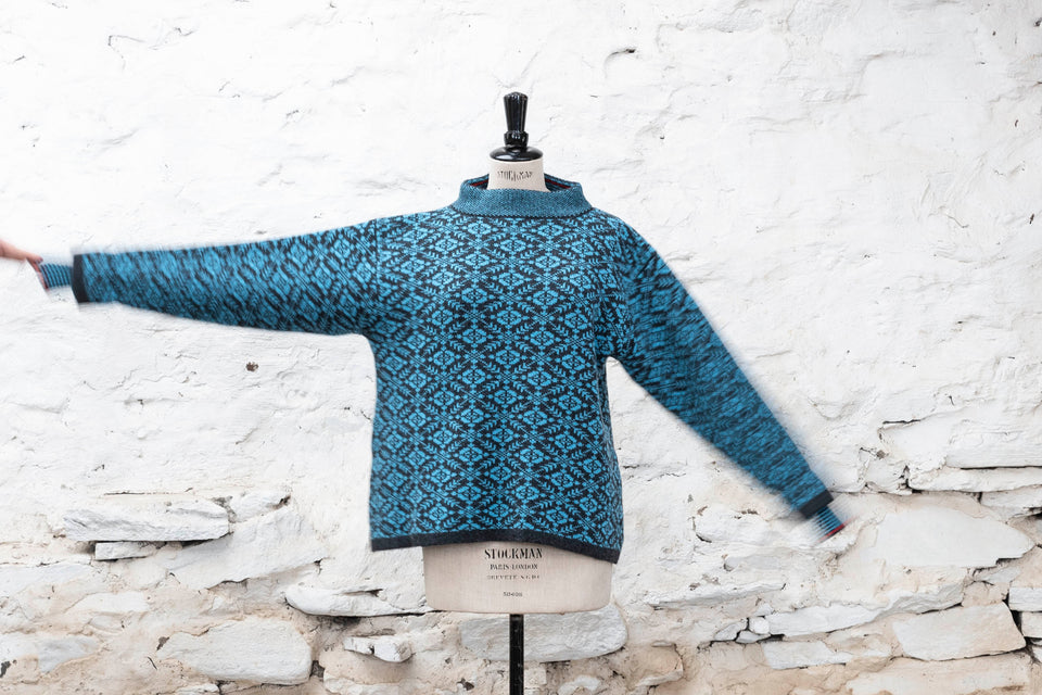 Fair Isle jumper. All over nordic inspired pattern with striped inset cuff and stand up neck. Shown in turquoise and charcoal with dark scarlet trim on a vintage mannequin, in a white washed Shetland shed.