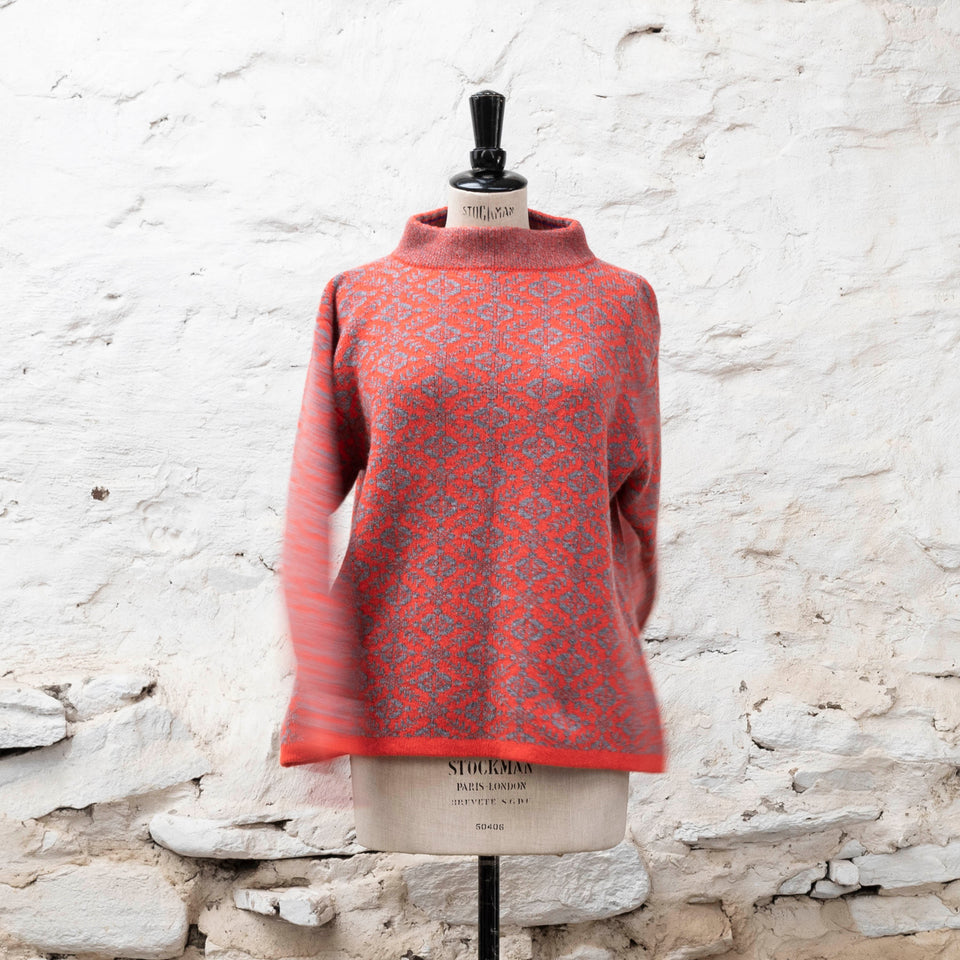 Modern Fair Isle sweater, made in Shetland. All over pattern with striped inset cuff and stand up neck. Shown in coral and grey on a vintage mannequin, with arms twisting, against a whitewashed rustic stone wall.