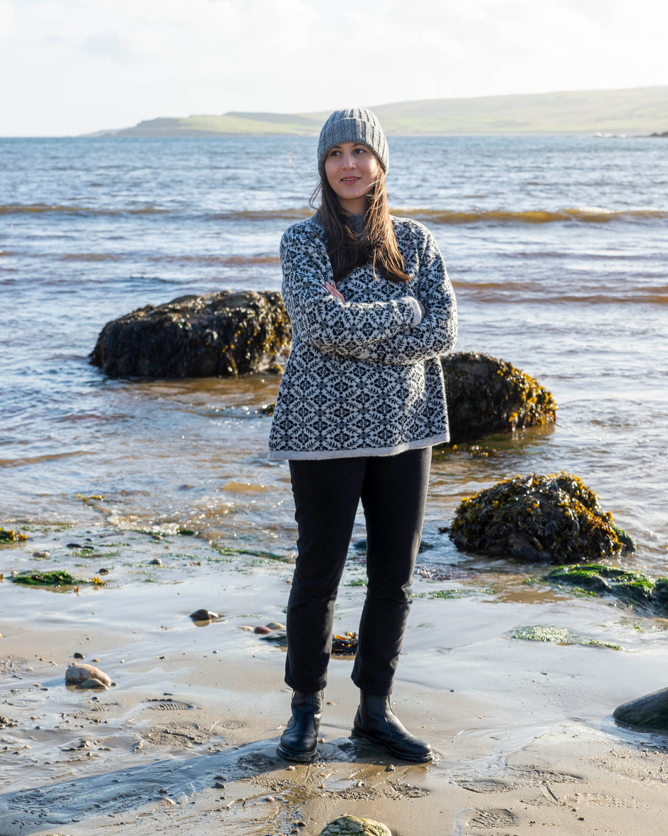 Fair Isle jumper in charcoal and pale grey. A woman with olive skin and long dark hair stands on the beach at Hoswick, Shetland. She is wearing a chunky grey beanie hat, the Fair Isle jumper in charcoal and pale grey and black jeans.