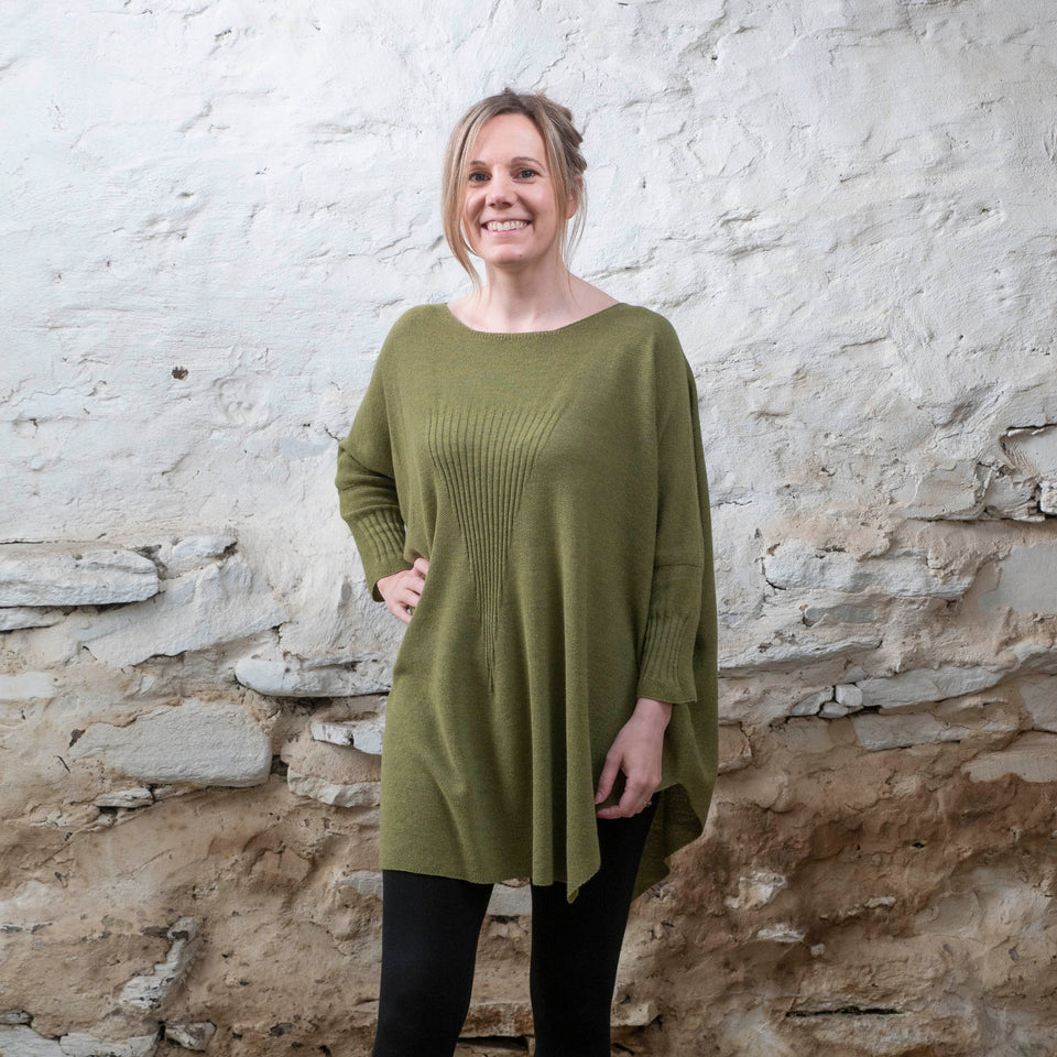 A white woman stands in a rustic stone shed. Her lightbrown hair is tied back and she looks straight at the camera and smiles. She wears a loose mossy green shetland jumper in a contemporary boxy style over black leggings.
